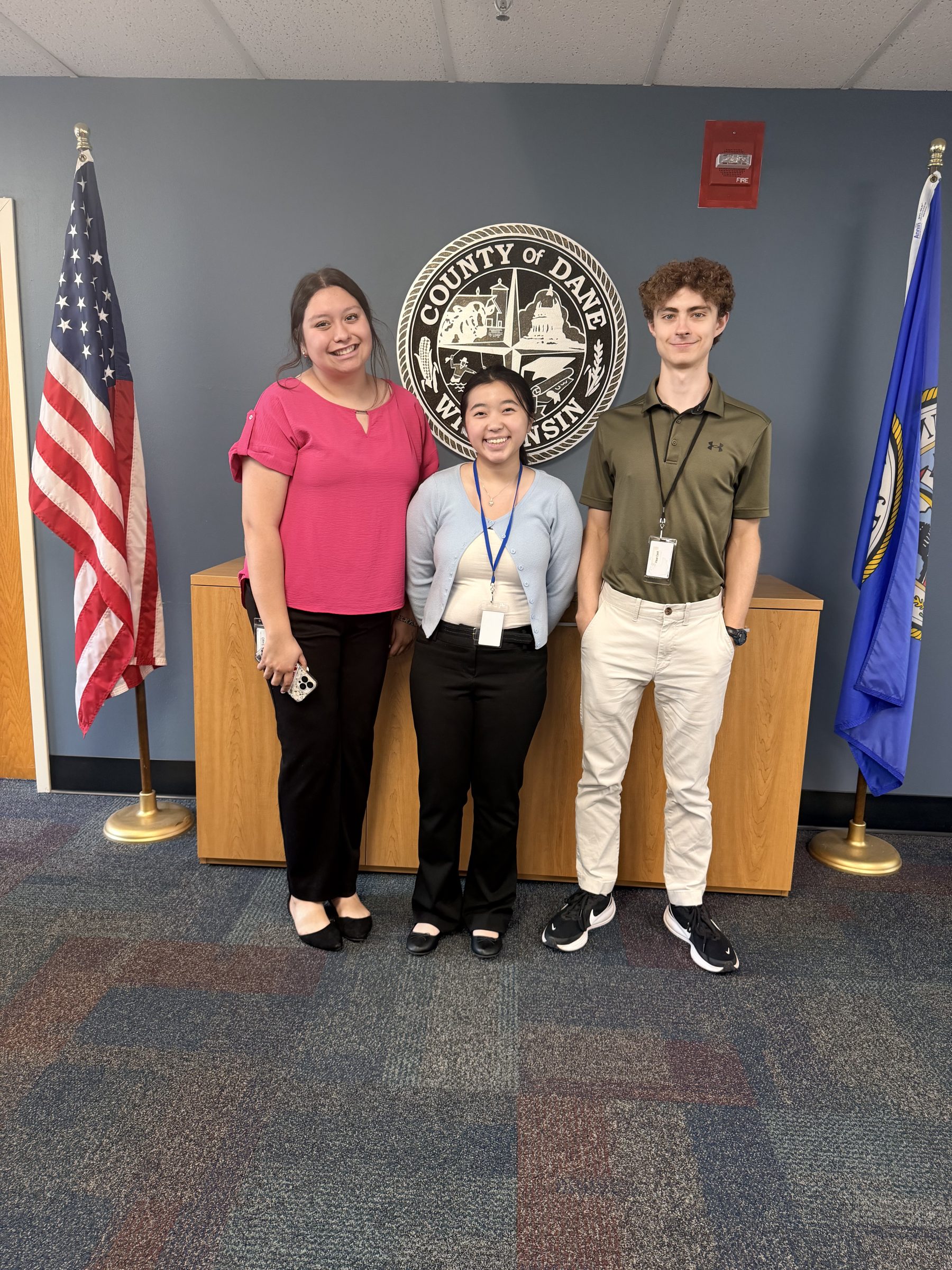 Three youths stand in front of the County Seal and pose side by side near county and us flags