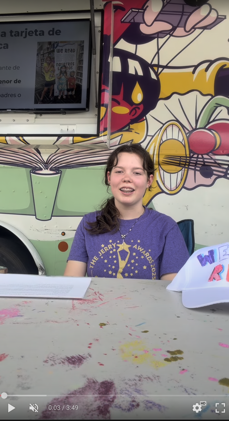 One young lady is staring the camera sitting in a chair with the Library Dream bus behind her